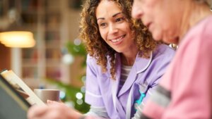 A senior woman wearing a pink shirt looking at a photo album with her female nursing assistant while smiling