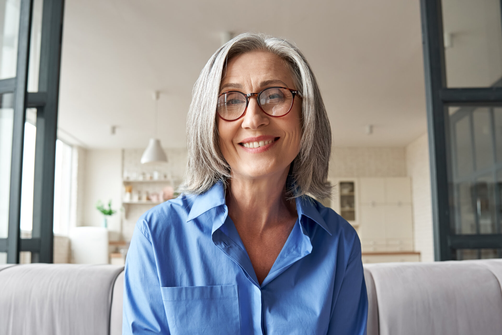 A senior woman with grey hair and glasses sitting on a grey couch while smiling